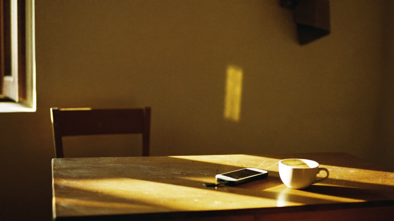 Mesa de madeira vazia num cômodo silencioso de manhã, com um celular esquecido de lado, uma xícara de café já fria e uma cadeira afastada