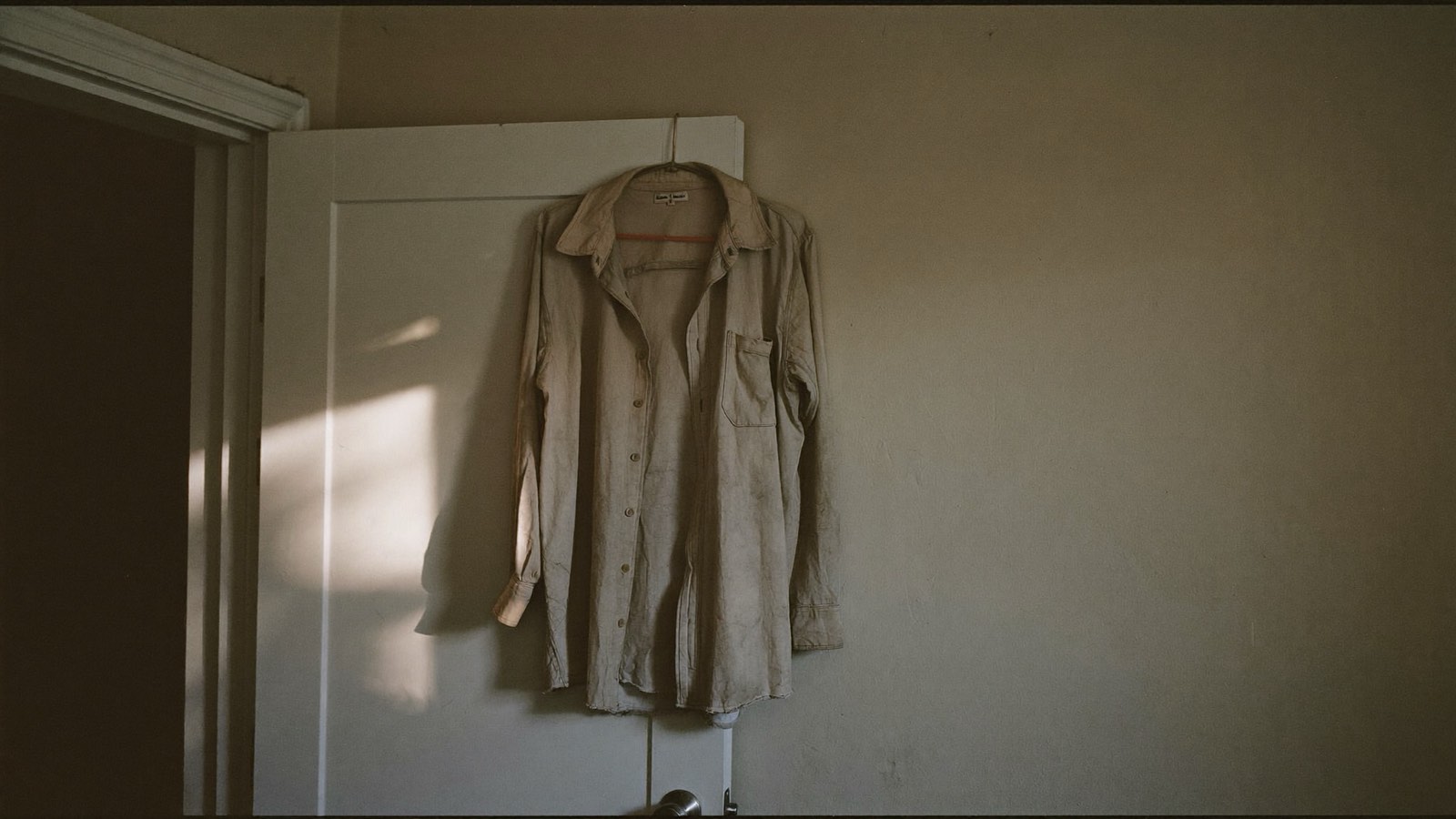 Camisa de trabalho gasta pendurada num gancho atrás da porta de um quarto, com a luz fraca de fim de tarde atravessando o cômodo